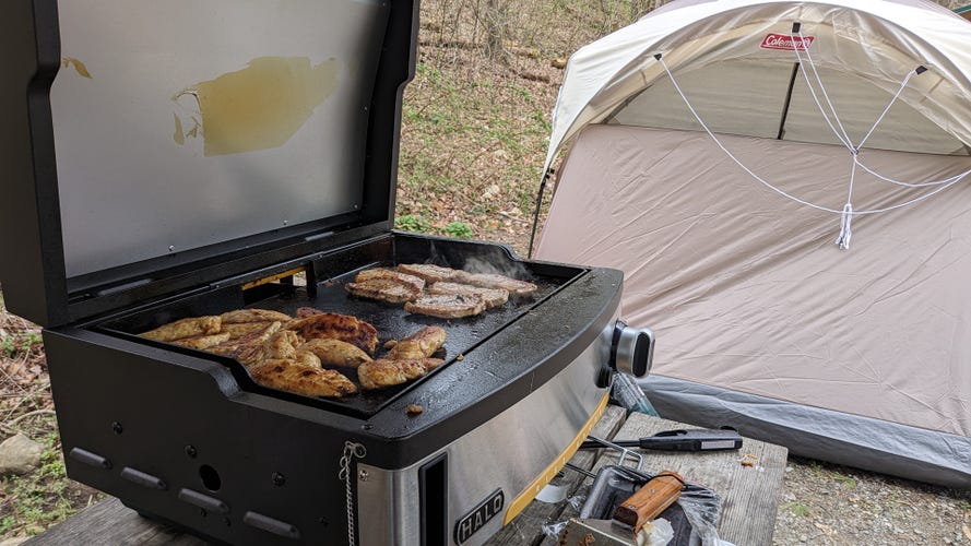 A tabletop griddle with chicken and pork cooking on it near a tent