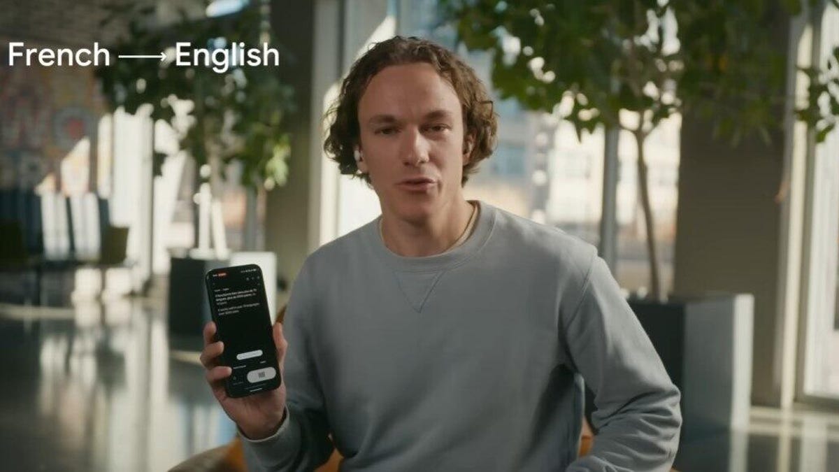 A man in a gray sweater holds up a phone that's doing live translation of French to English language. He's wearing earbuds.