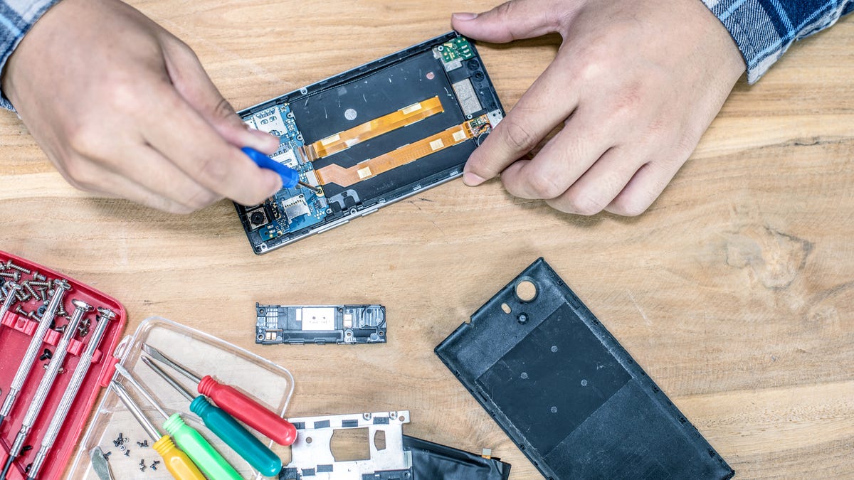 Overhead shot of hands working on an opened smartphone. The phone's innards are visible, as is a set of tools, including a selection of small screwdrivers.