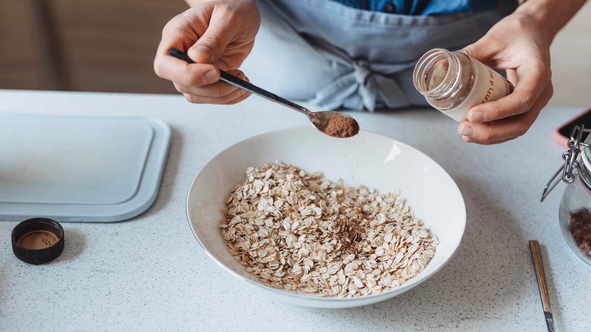 A close-up of cinnamon being sprinkled in a white bowl of oats with a spoon.