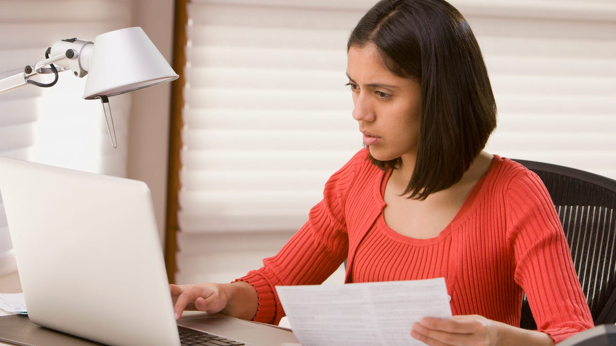 A person frowns at a laptop while holding paper documents.