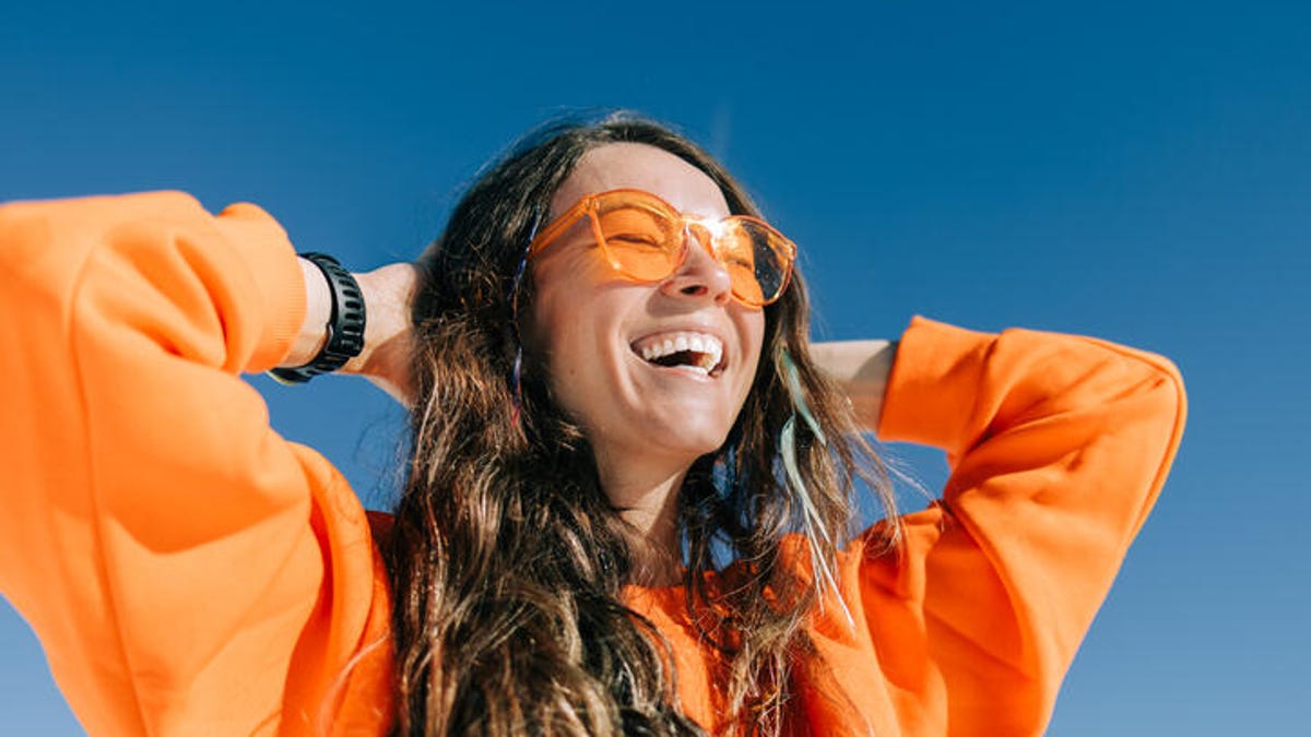 Young woman wearing an orange sweatshirt and sunglasses smiling while standing in the sun.