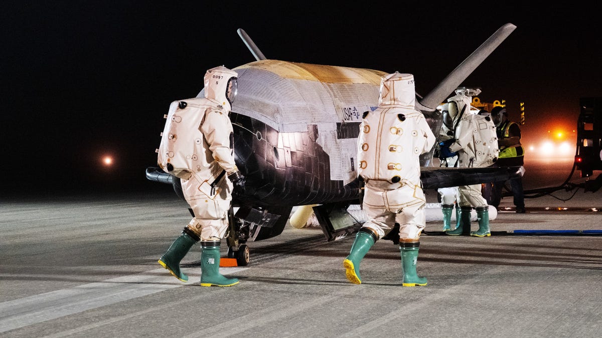 X-37B space plane resembling a small space shuttle on the runway with several workers in safety suits and helments checking it out.