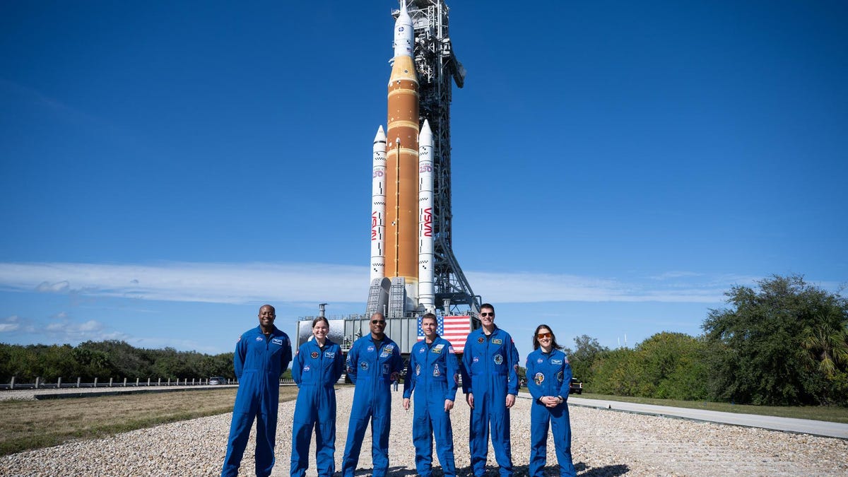 Six people, clad in blue NASA jumpsuits, stand in front of the SLS rocket system