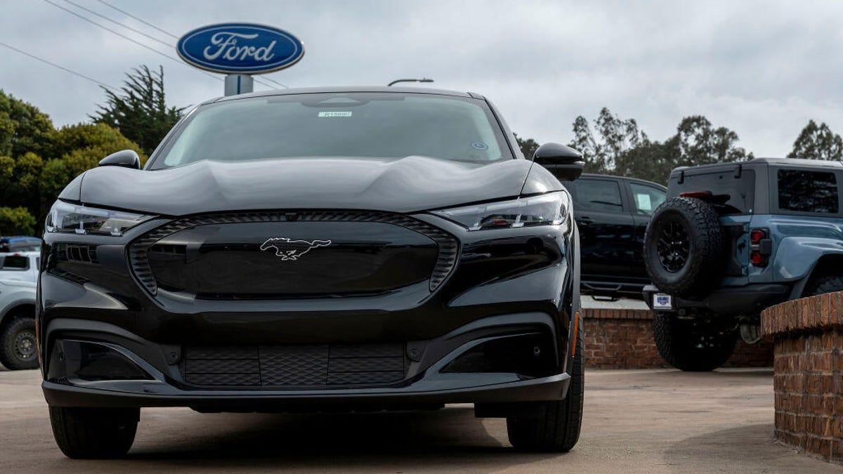 A Ford Mustang Mach-E sits in a dealership lot, with a Ford sign in the background.