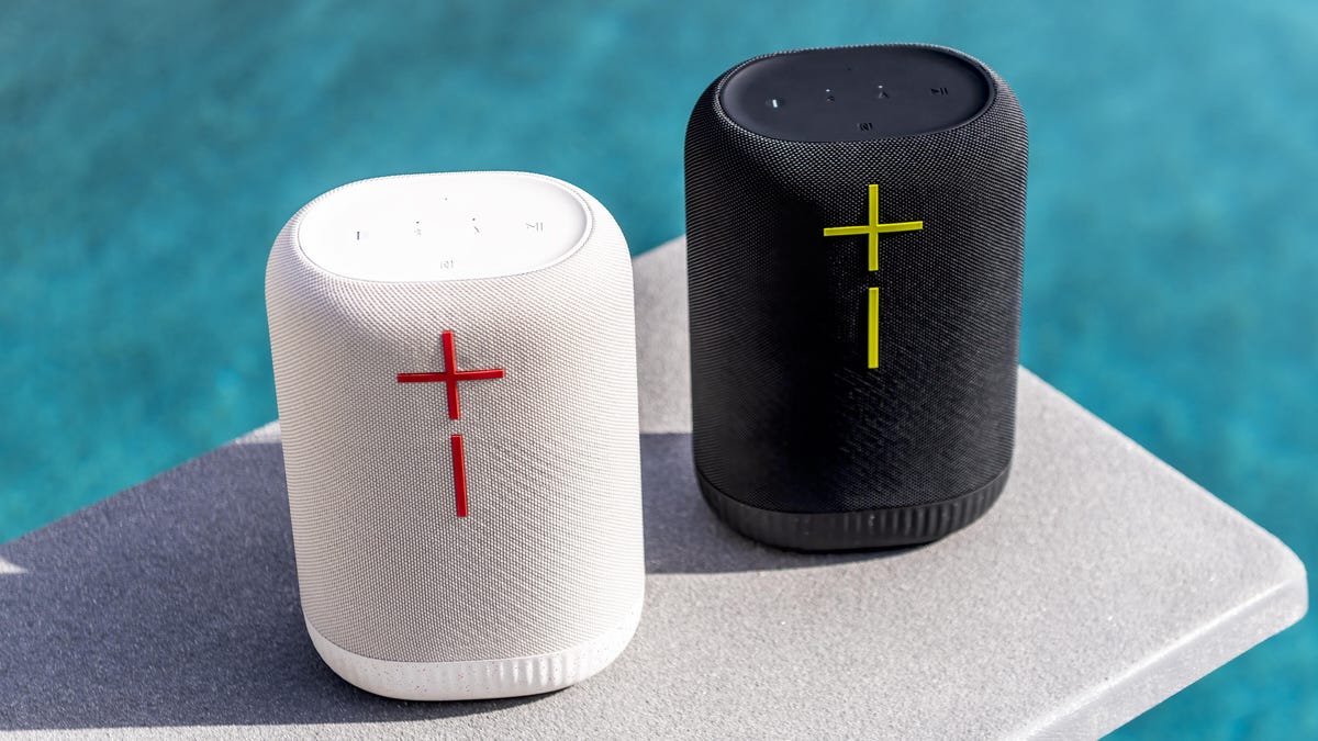 One black and one white UE Epicboom Bluetooth speaker, sitting on a diving board over a swimming pool