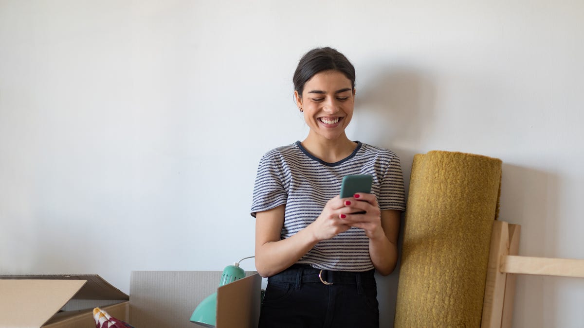 A person smiles at their phone next to a rolled-up rug and moving box.