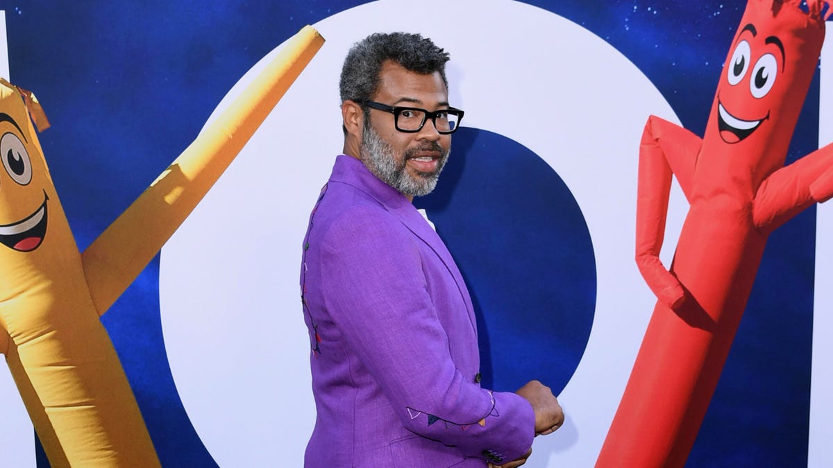 Jordan Peele in a purple suit in front of a poster for Nope