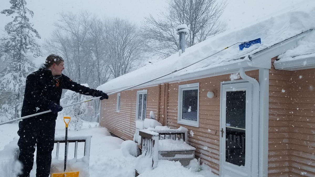 During a snowstorm, a man uses a roof rake to clear snow from the roof of a house.
