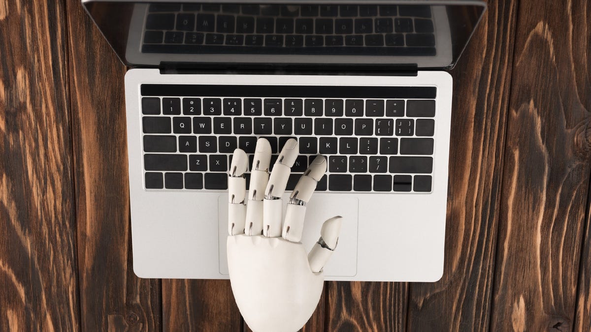 A robotic hand reaches over a laptop keyboard, poised to write.