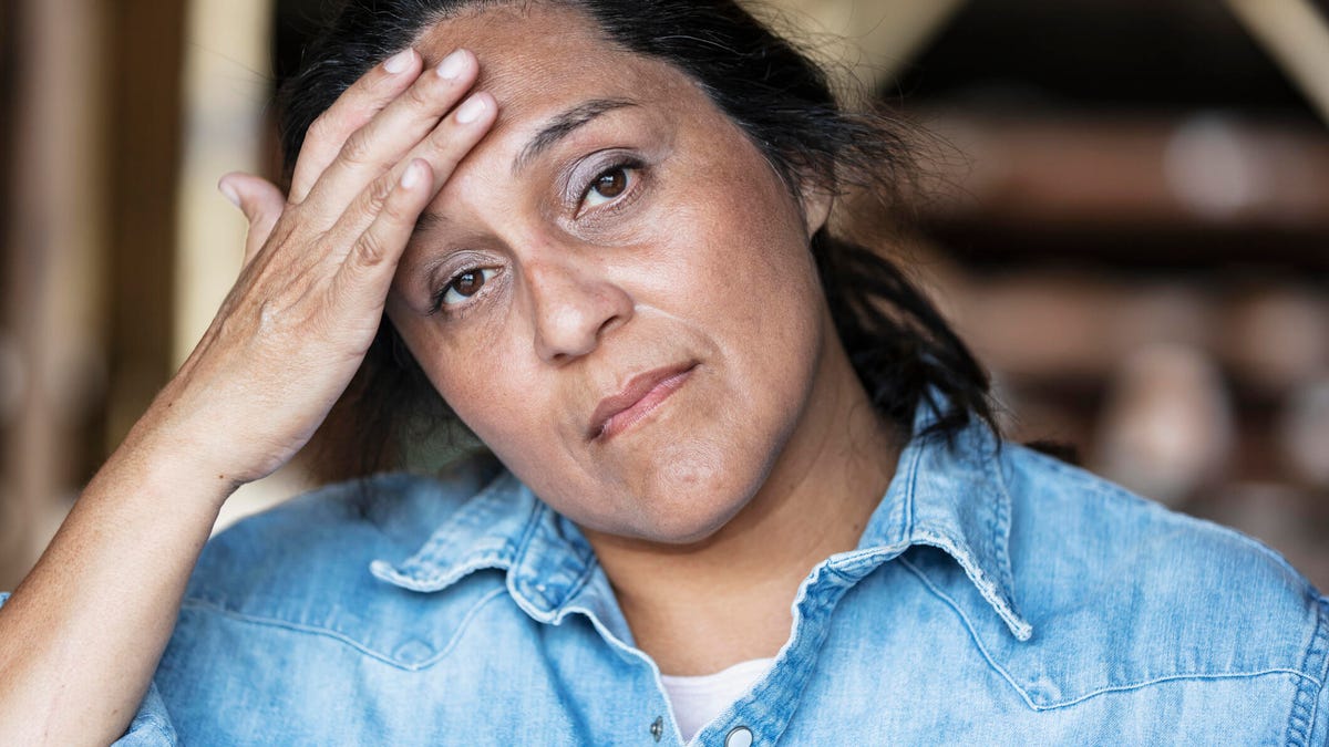 A woman in a denim shirt with pulled-back long black hair placing a hand on her forehead.