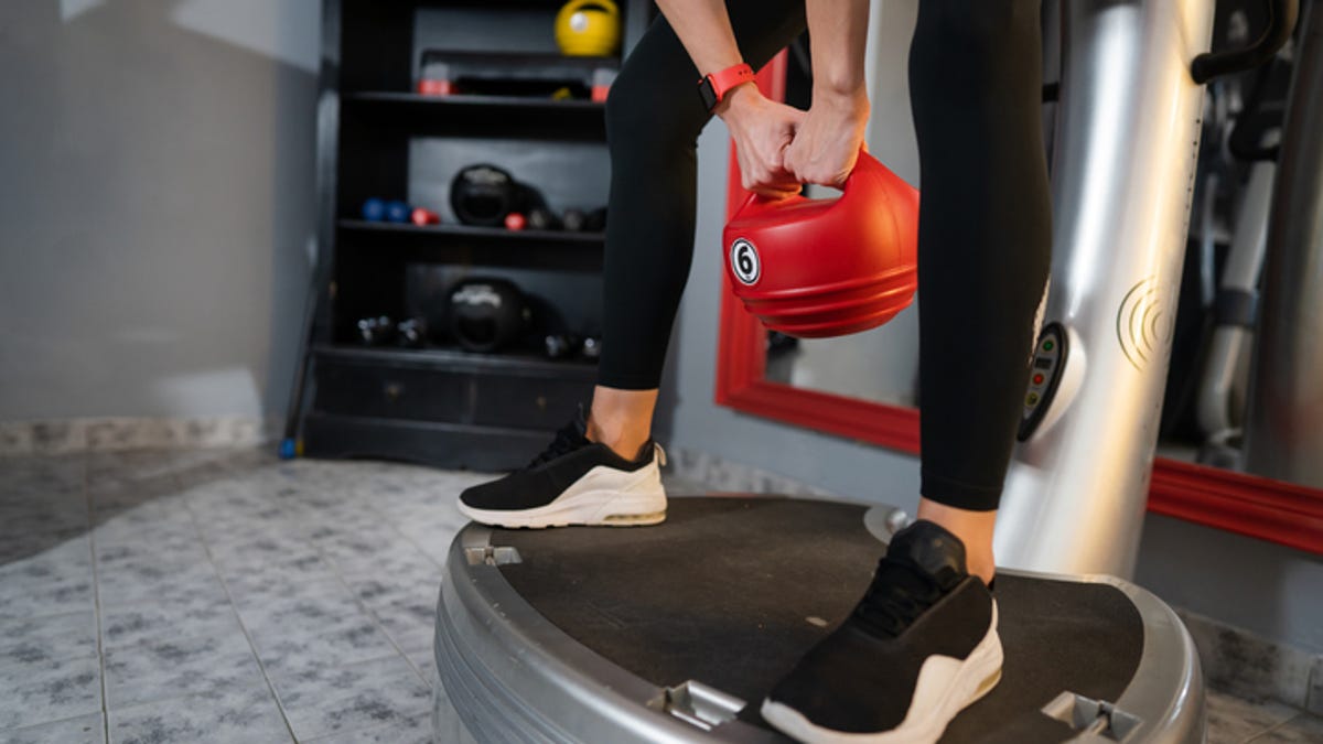 A close-up of a person wearing grey leggings and sneakers standing on a vibration plate in a living room with a mustard yellow couch.