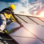 A solar panel installer places a solar panel on top of a house as the sun sets.