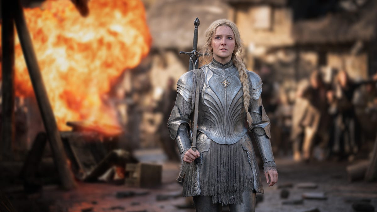 A young Galadriel standing in silver armour with a fiery explosion behind her in a village