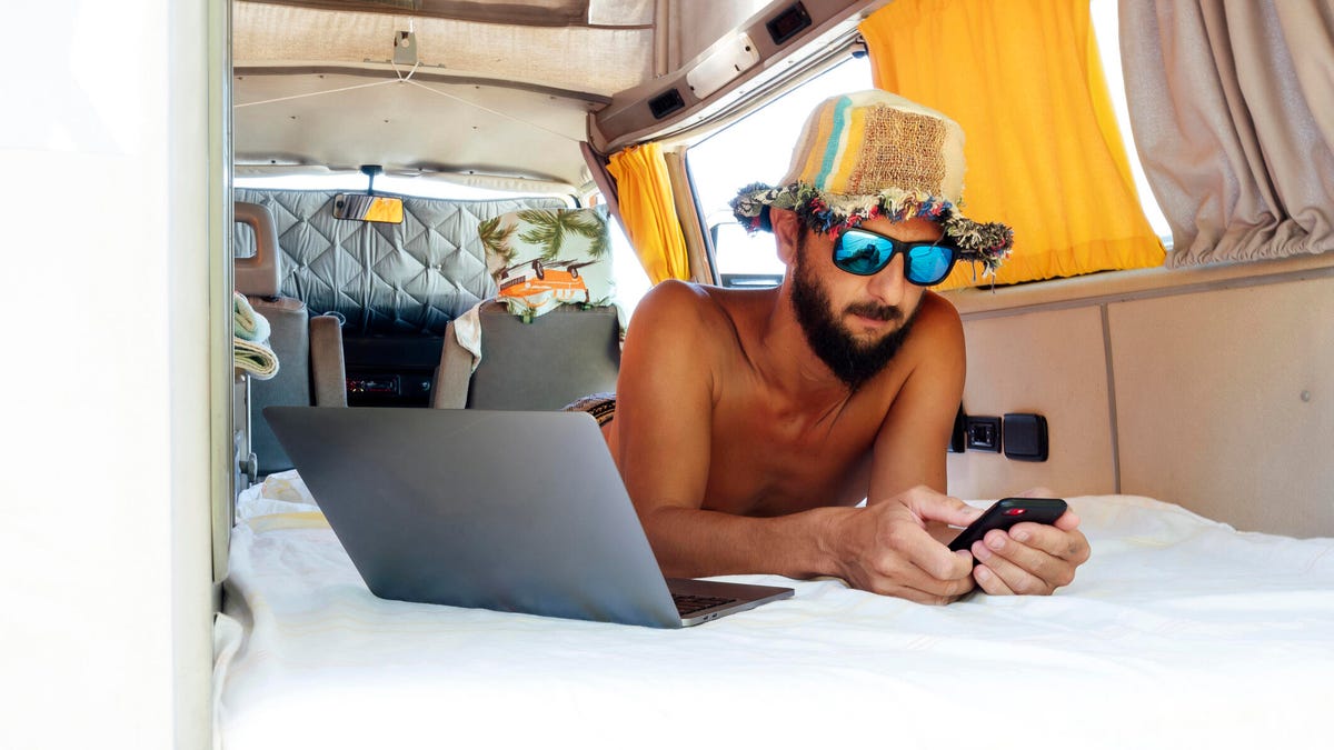 A man lies in a van using his phone with a laptop beside him.