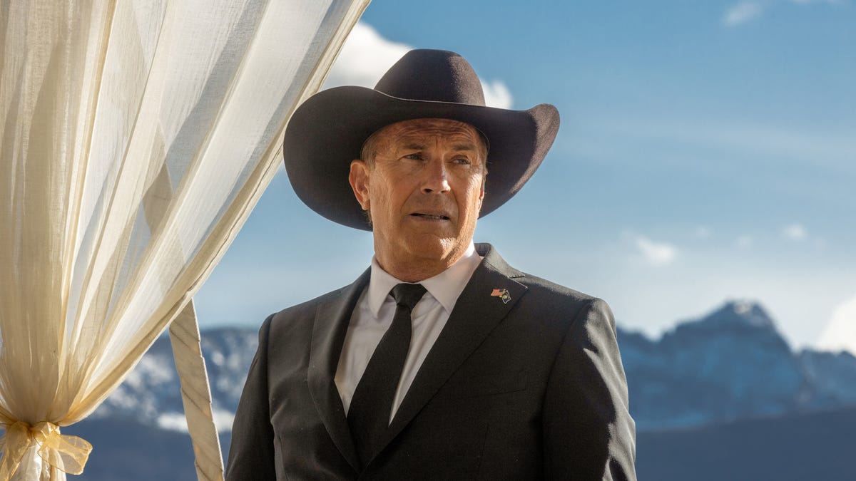 John Dutton, played by Kevin Coster, stands in his cowboy hat and suit with Montana mountains in the background.