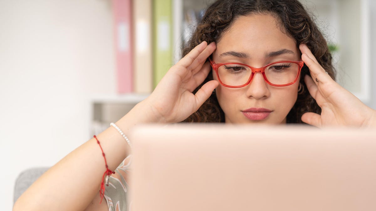 A woman at home touches her temples while looking at a beige laptop.