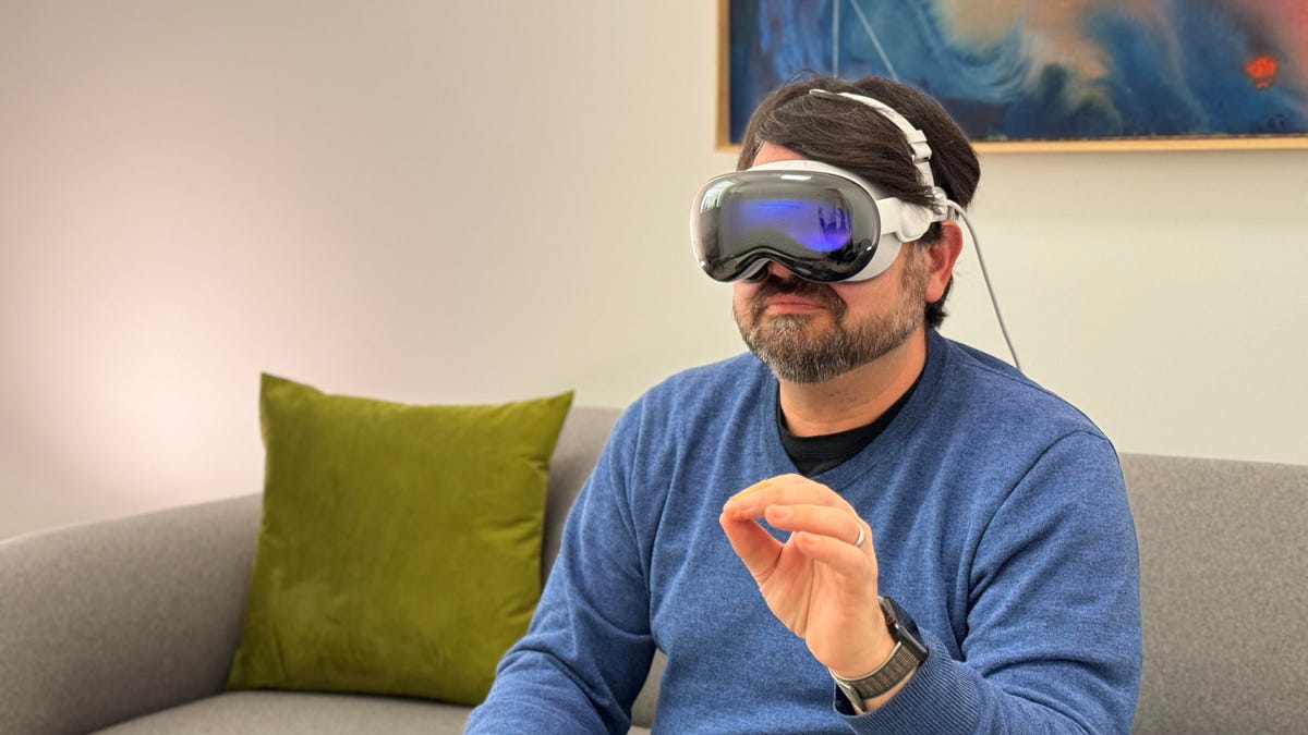 A man in a blue sweater on a green sofa pinching his fingers and wearing an Apple Vision Pro headset