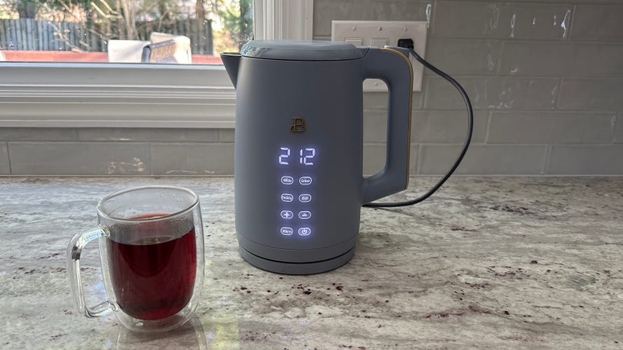 The Beautiful by Drew Barrymore One-Touch Electric Kettle sits on a granite countertop beside an inviting cup of tea.