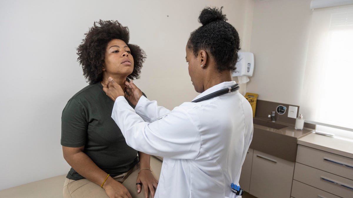 A doctor with black hair in a bun examining a patient's neck on an exam table in their office.