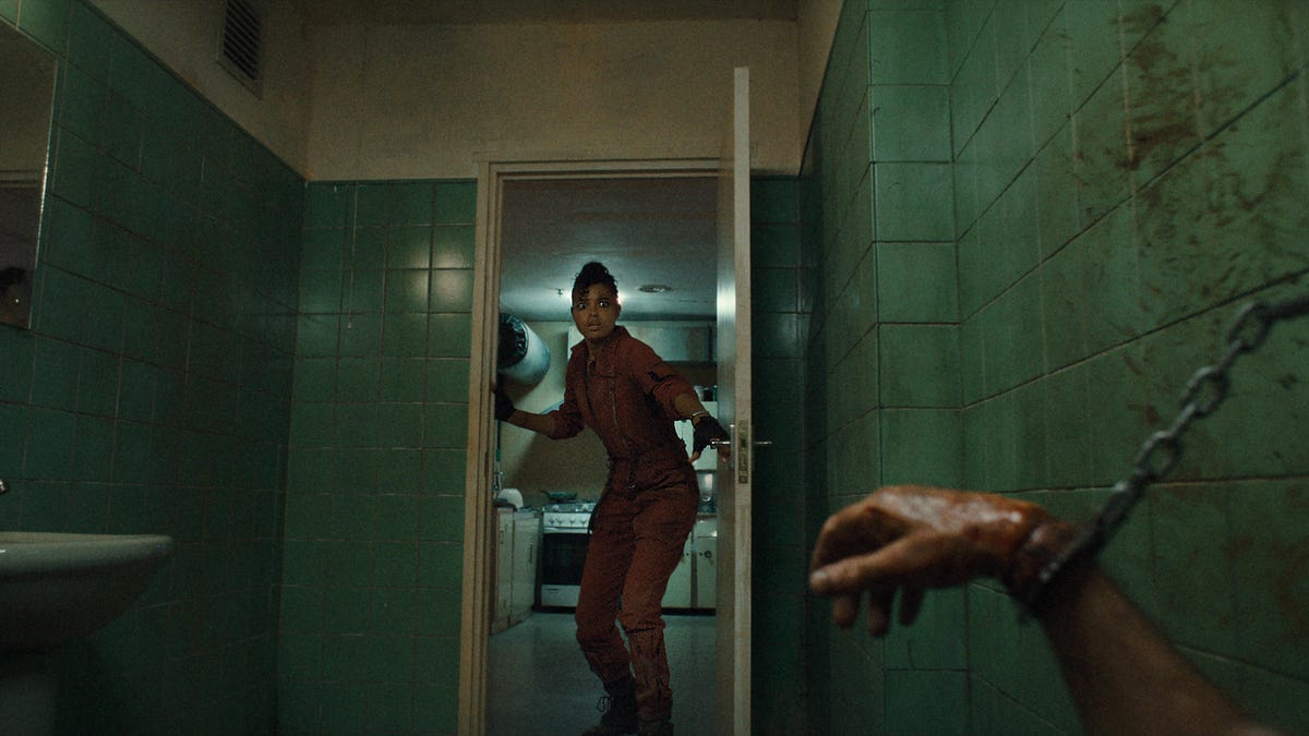 A woman opens a door where a person is chained to the wall by their wrist.